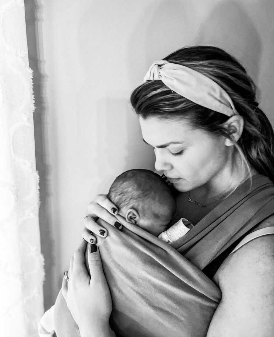 New mom holding her baby on a baby carrier wrap kissing the baby's head