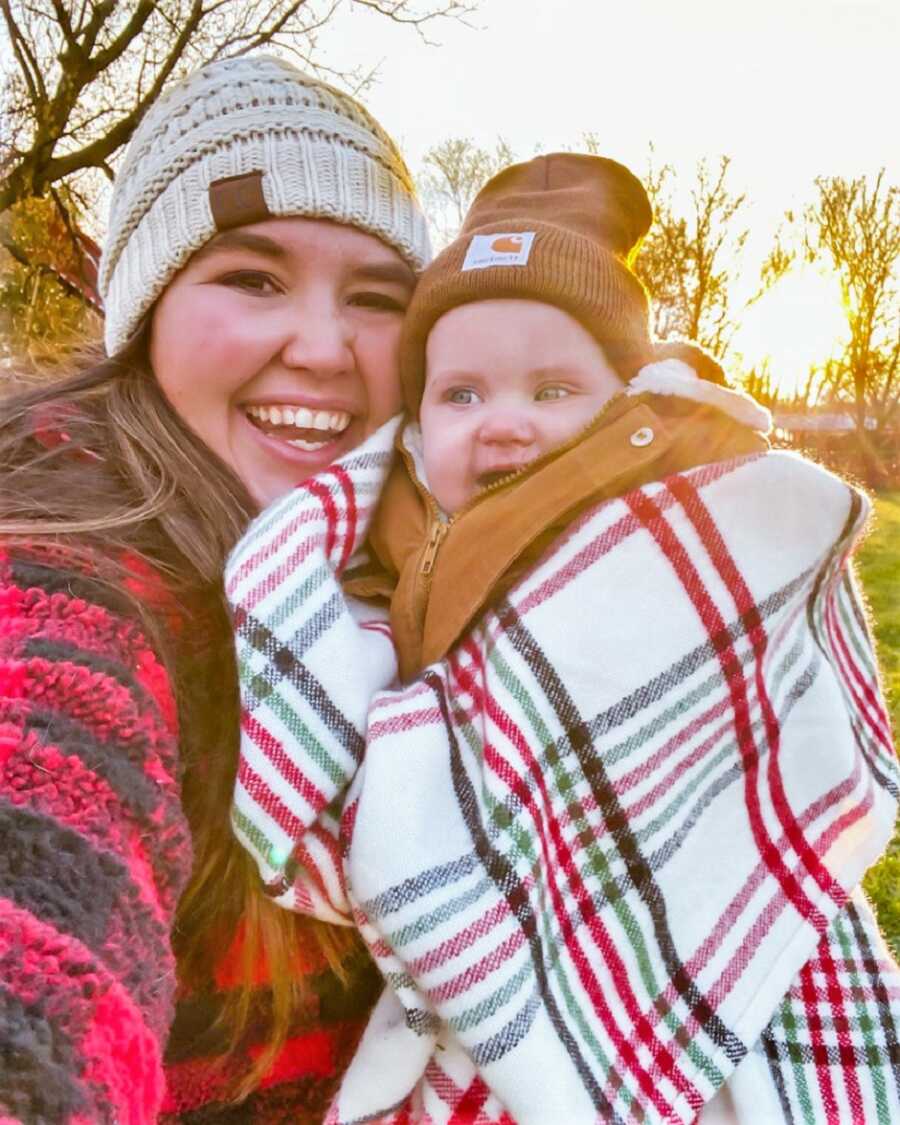 Mom and daughter wearing hats and plaid sweaters smiling with the sunset in the background