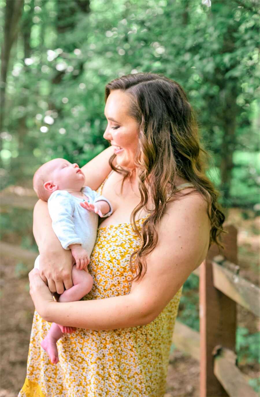 mom wearing a yellow floral dress holding baby girl in her arms and looking at her while smiling 