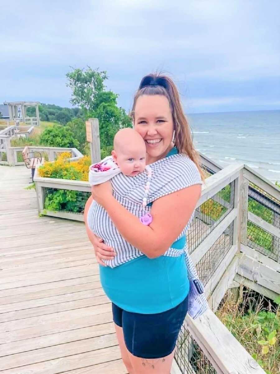 Mom wearing a navy striped baby carrier wrap with her daughter at the beach smiling 