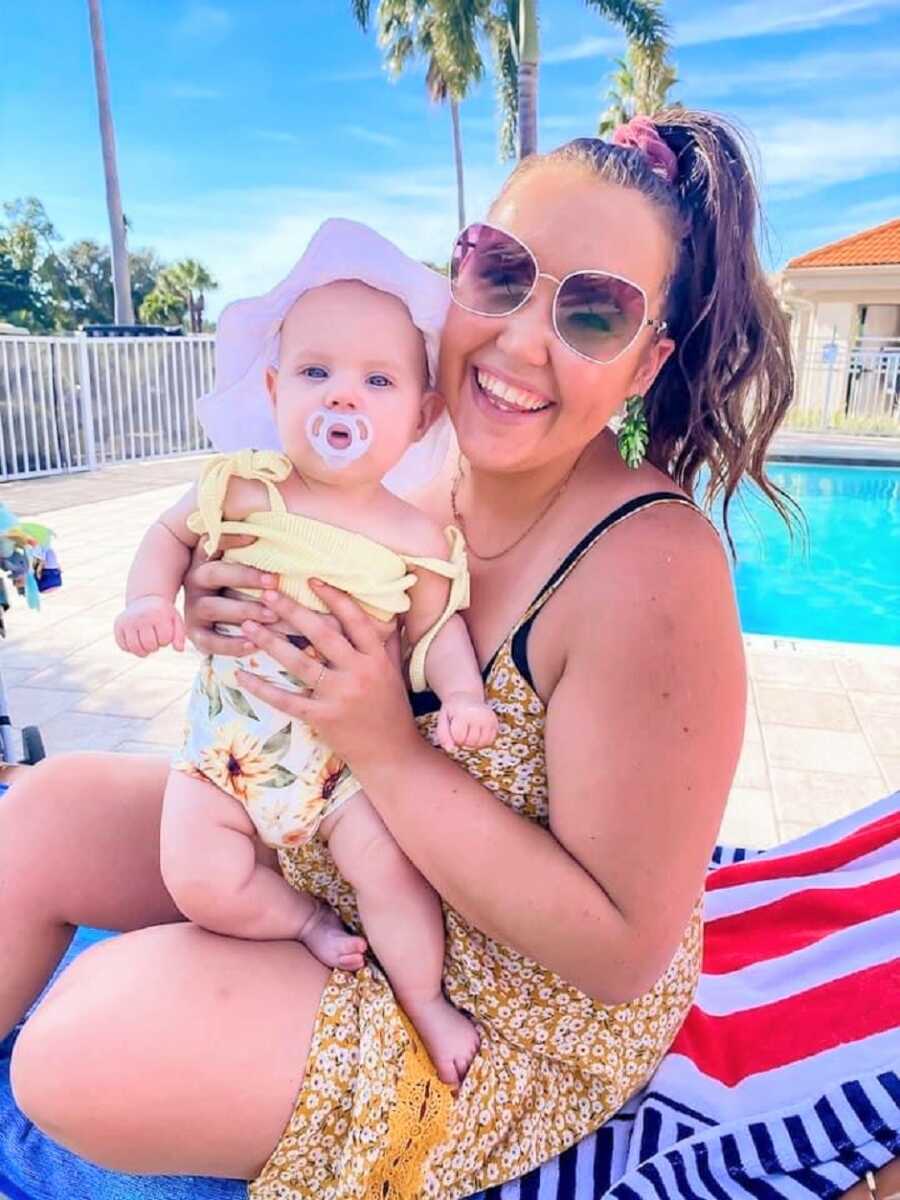 Mom and baby daughter in bathing suits sitting in front of the pool while baby has a pacifier in her mouth and a big white hat