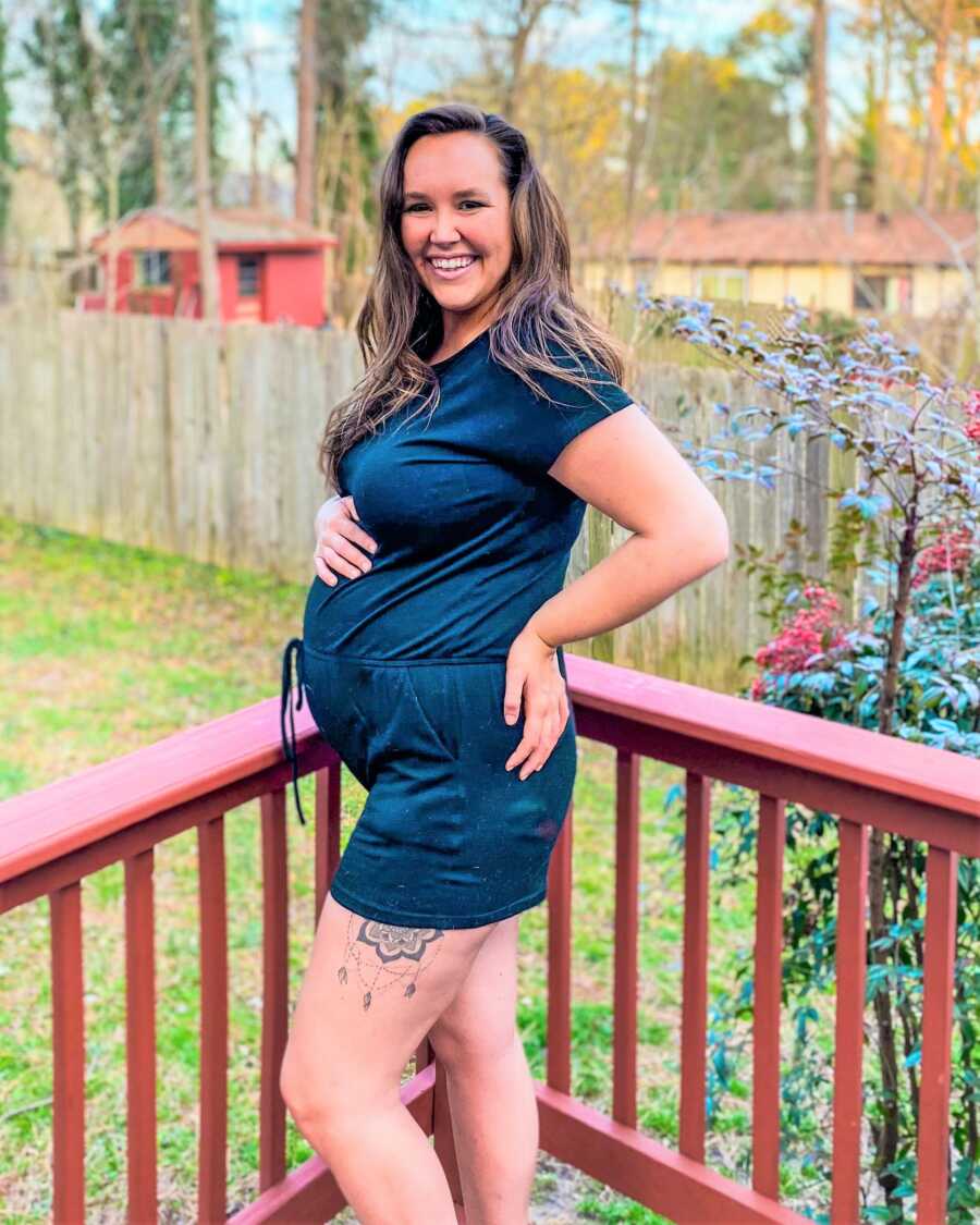 Pregnant woman wearing a blue jumper holding her belly and smiling at the camera while standing on a porch red porch 