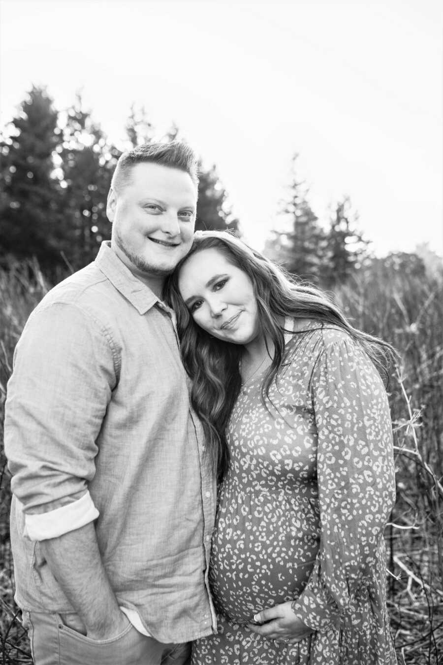 black and white image of pregnant couple in a grass field with mom-to-be holding belly and wearing a long sleeve dress 