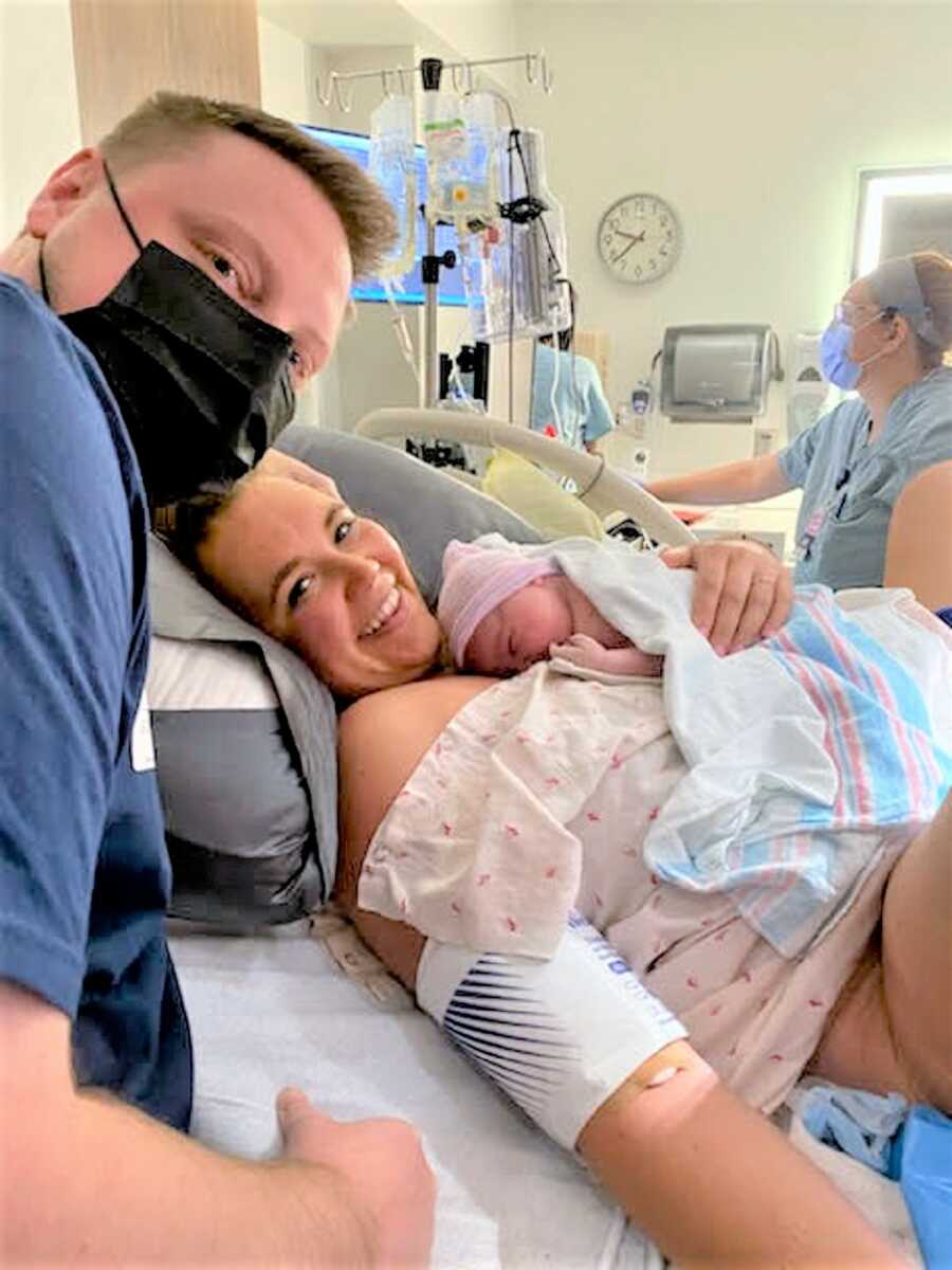Mother holding newborn baby at the hospital next to dad wearing a mask over his mouth and nose 