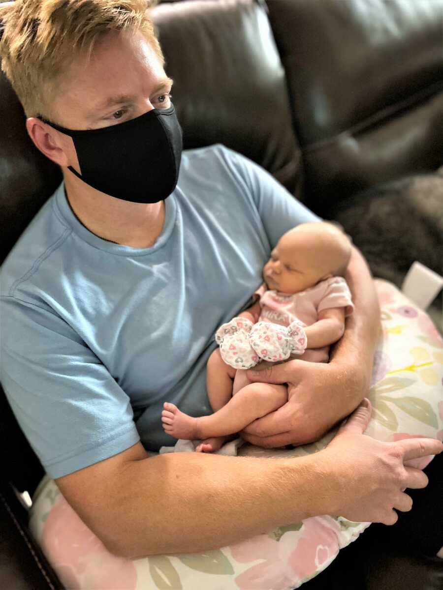 New father sitting on a black couch holding newborn baby girl on a pillow and wearing a black mask because of COVID