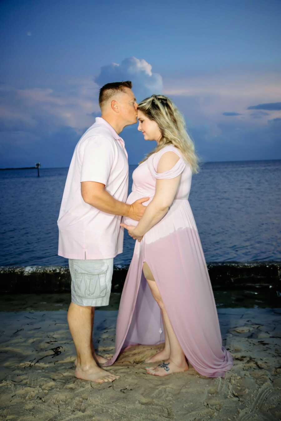 husband kisses pregnant wife in the forehead while holding pregnant belly in front of the beach wearing a pink shirt and a pink dress