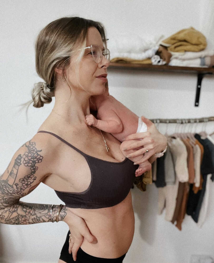 Postpartum mom holding her newborn baby on her chest, wearing a bra and underwear with a clothing rack in the back 