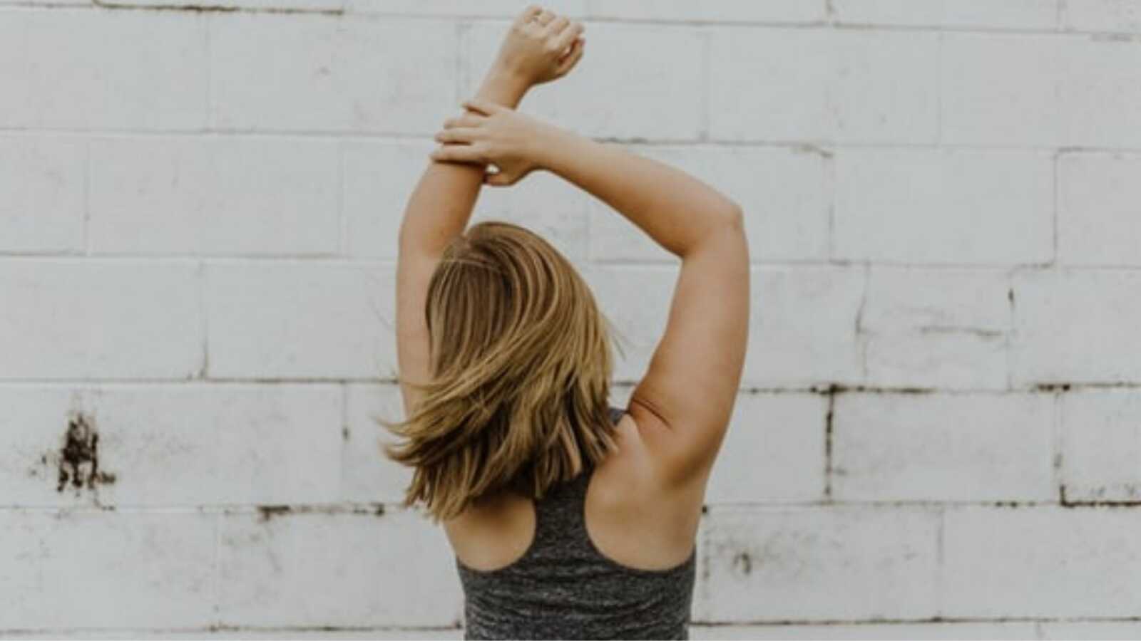 Woman dances in front a white brick wall while wearing a workout tanktop