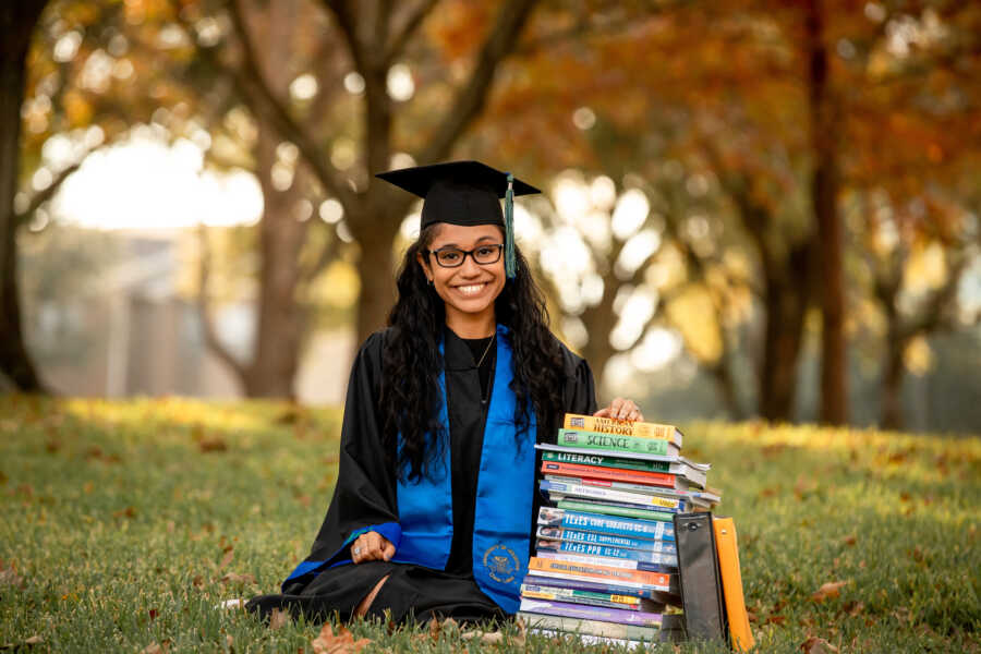 Young woman graduating from college takes a photo in her cap and gown while sitting next to a pile of her old textbooks