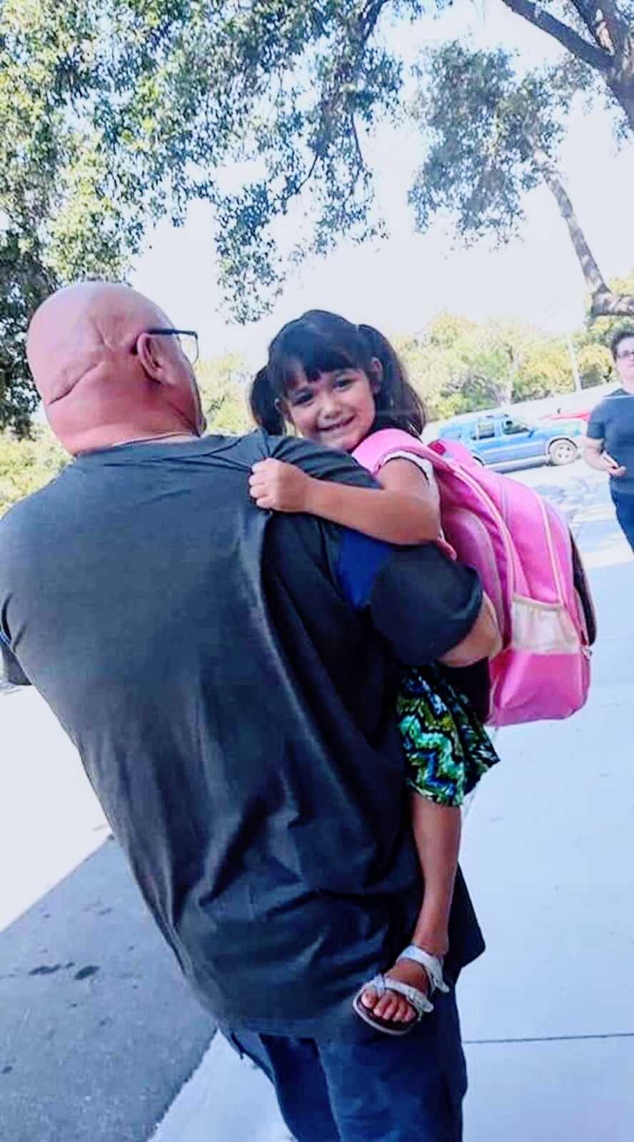 grandfather carries his granddaughter at his side while walking, she has a backpack on