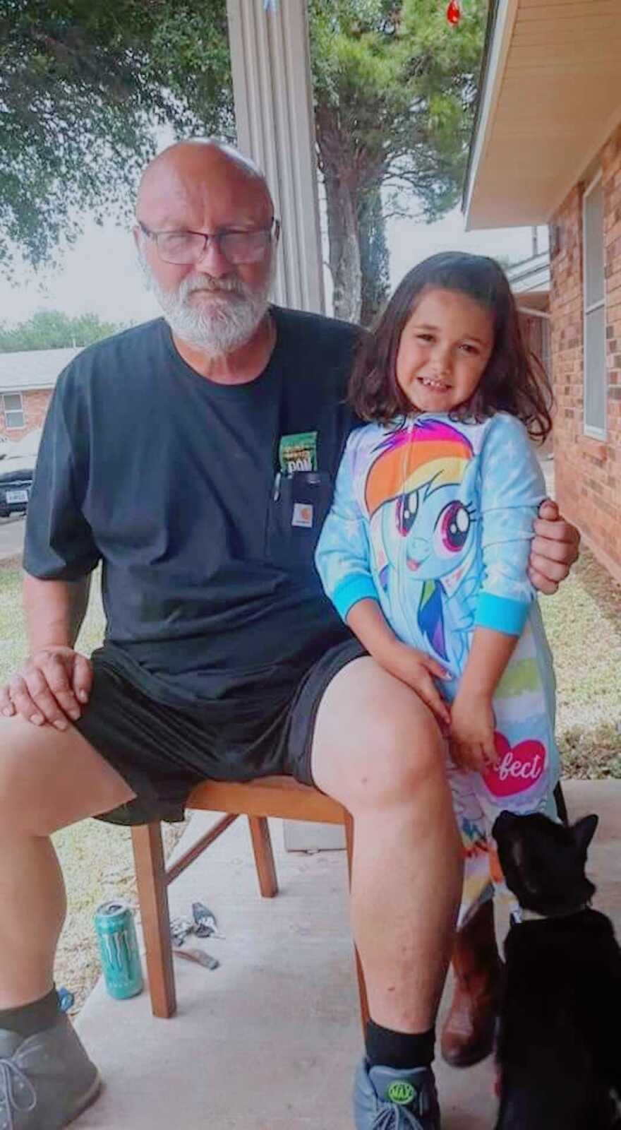 grandfather with granddaughter at his side, he is sitting in a chair while she stands next to him with an arm around her