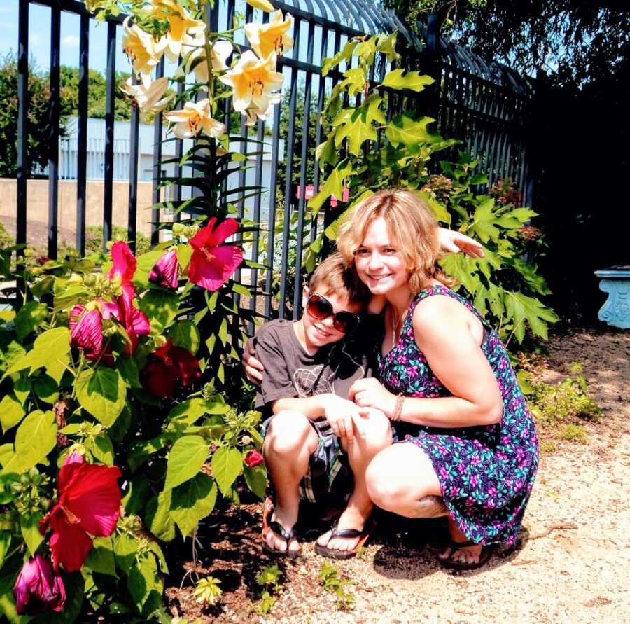 A mom and her young son sit in a garden near a fence