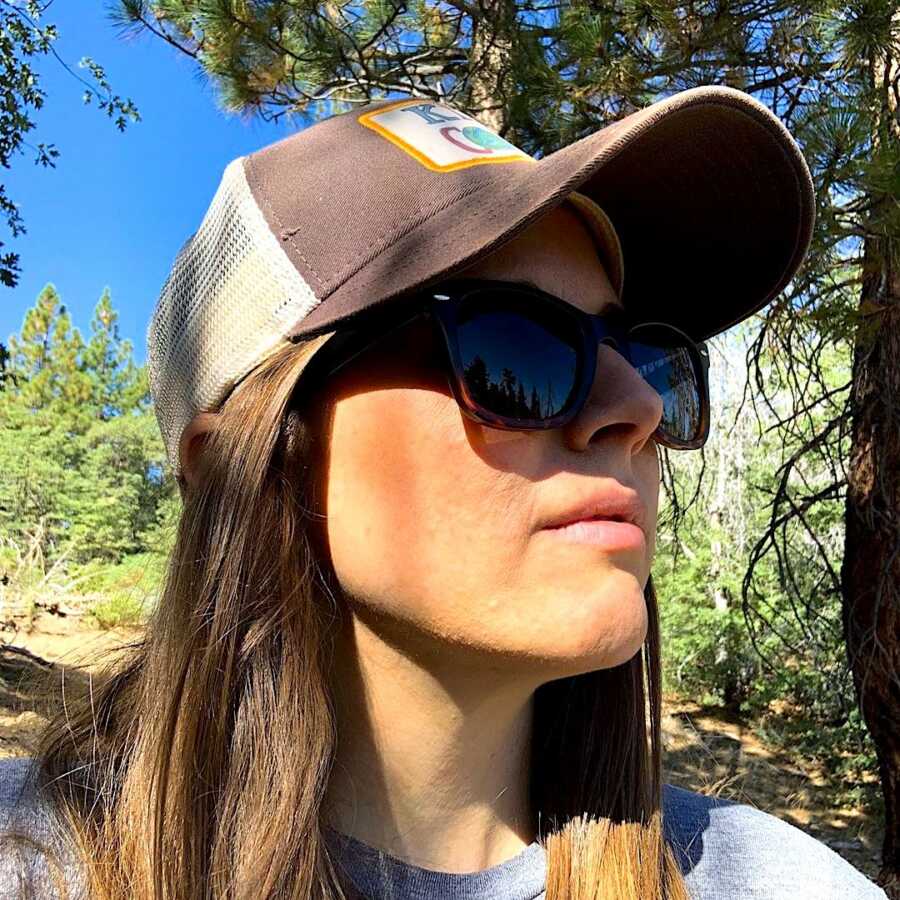 woman taking a selfie in the woods wearing a hat and sunglasses, looking to the right