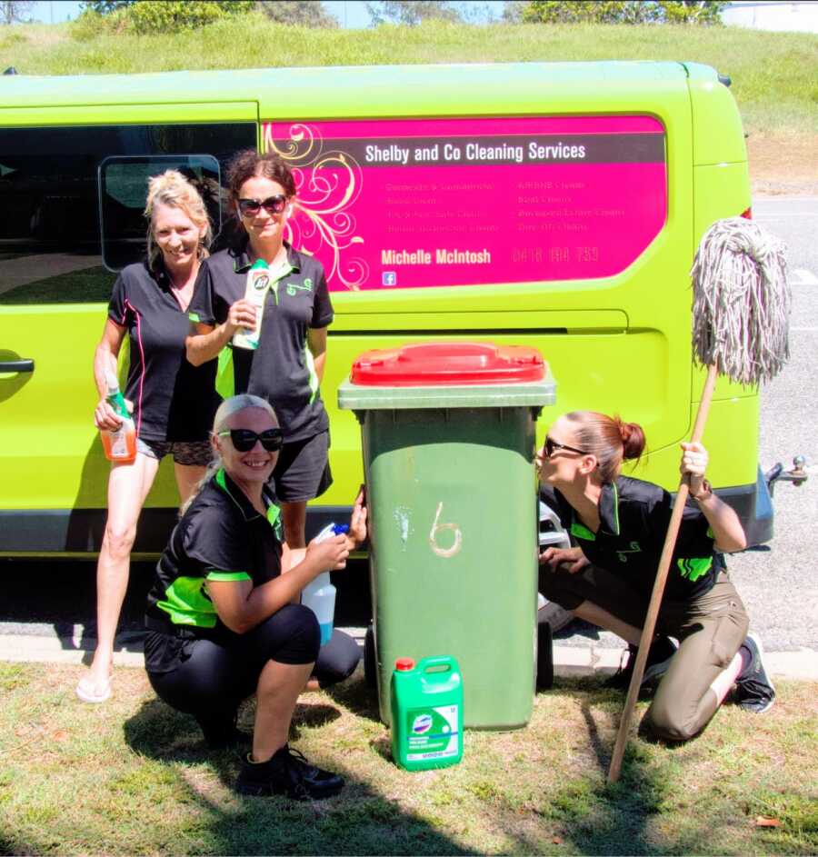 four women pose with a garbage can holding cleaning products in front of a cleaning services van