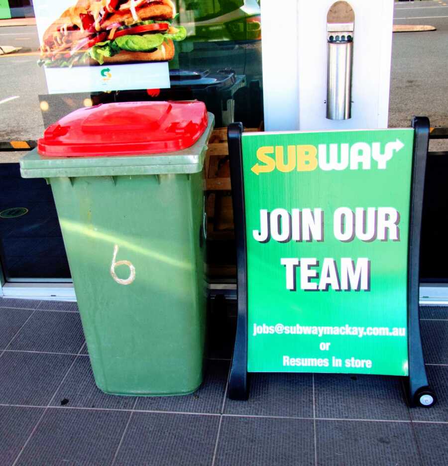 garbage can sits in front of a Subway restaurant next to a hiring sign