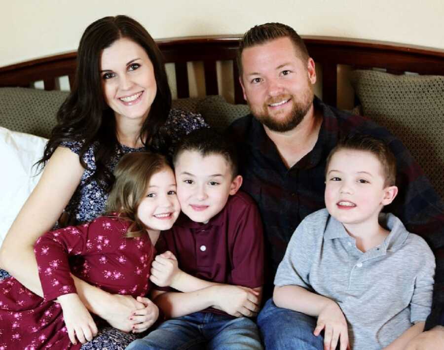 Couple takes family picture with three children sitting on their bed.