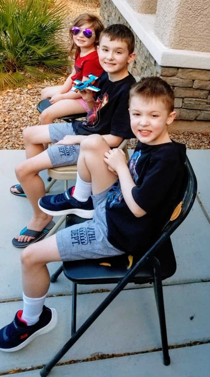 two boys with Duchenne's sit in folding chairs along with their sister, all of them are looking towards the camera, smiling