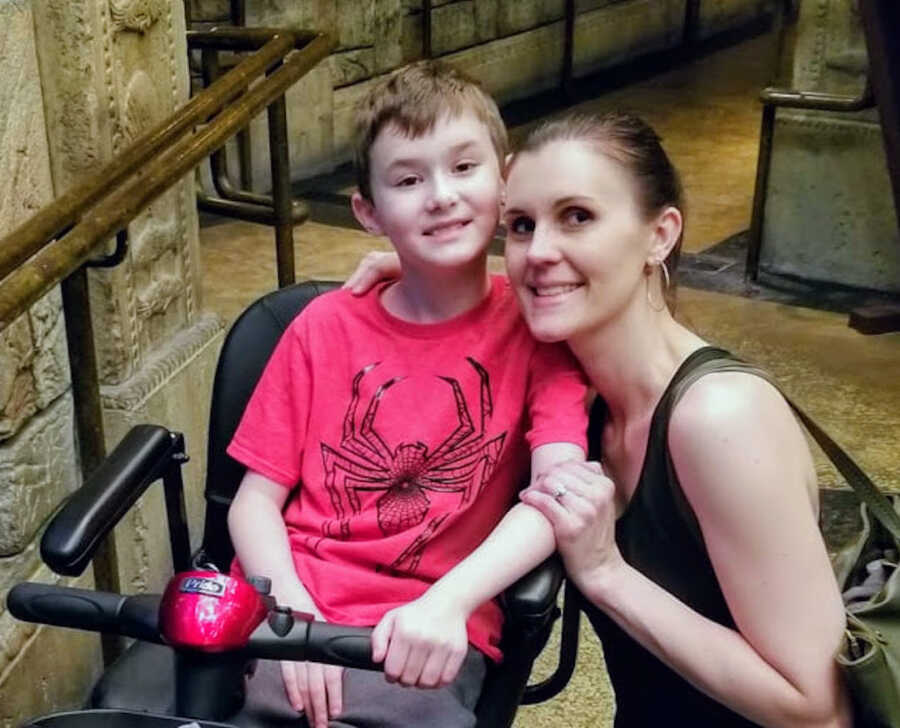 young boy with Duchenne's sits in his wheelchair while his mother bends towards him with an arm around him