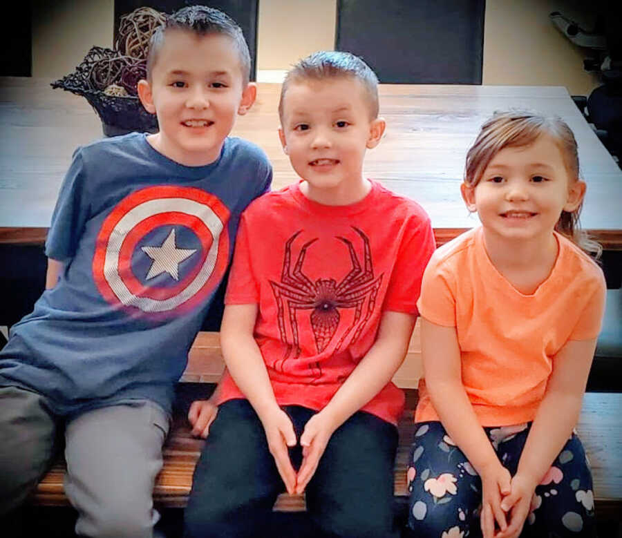 three siblings, two boys with Duchenne's and a girl sit at a table bench smiling