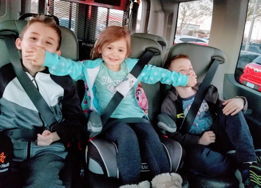 three siblings sit in the backseat of a van, the daughter is in the middle and has her hands over her two brother's mouths