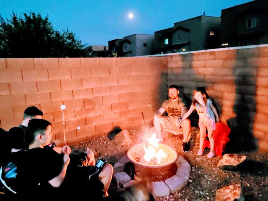 three children and their father gather around a fire pit at dusk and are roasting marshmallows
