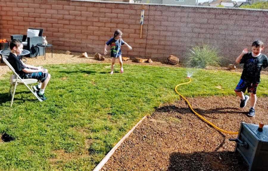 two young boys with Duchenne's play outside in a sprinkler with their sister, one of the boys is sitting in a chair