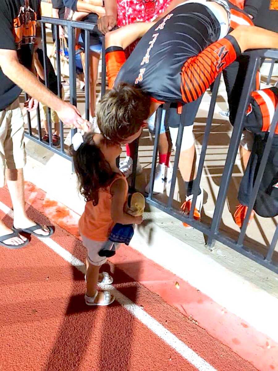 teenage son bends over fence after a sports game to give a young girl a kiss