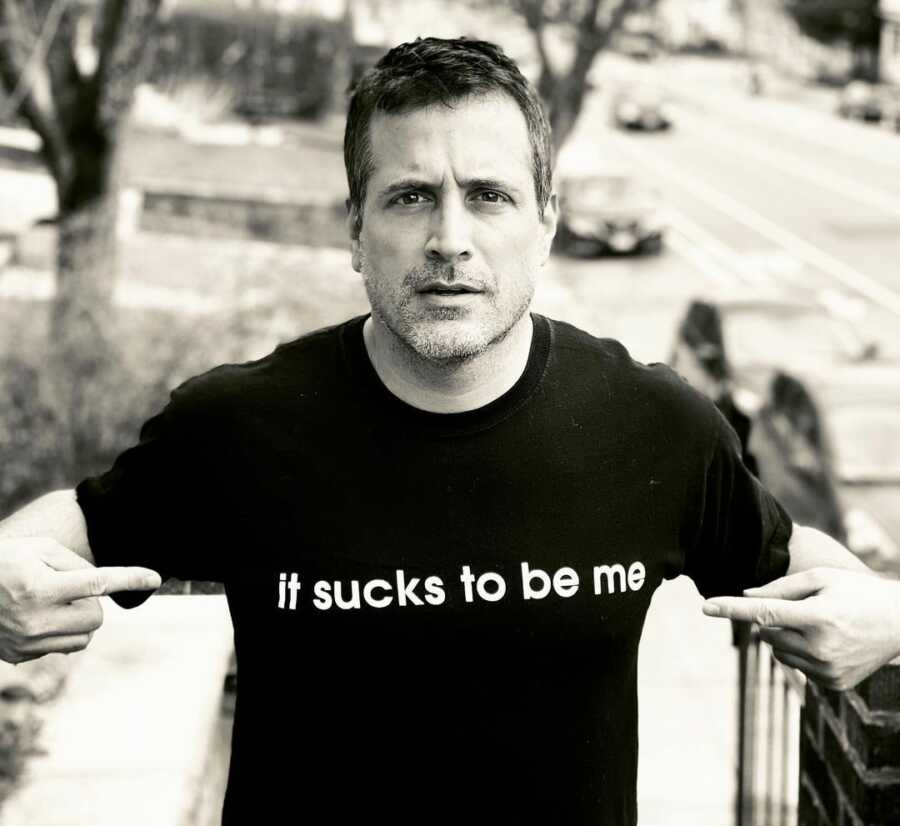 single father poses for camera wearing a graphic shirt that says "it sucks to be me" while pointing towards the shirt 