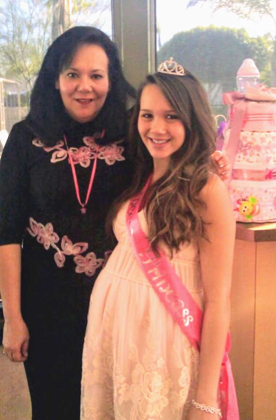 pregnant team mom poses with family member at her baby shower, she is wearing a sash and a crown
