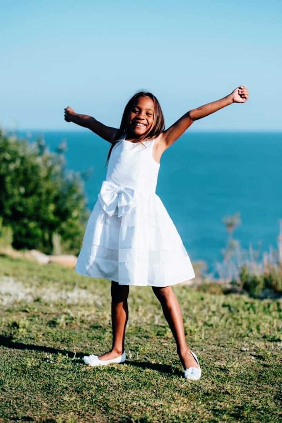 young girl poses outside by the water with her arms outstretched and a smile