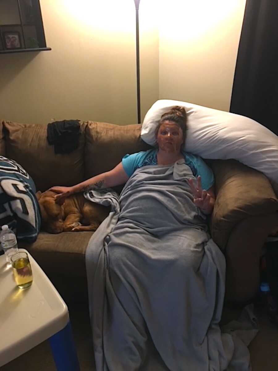 pregnant woman sits on couch with her dog at her side while shes covered in a blanket and wearing a face mask