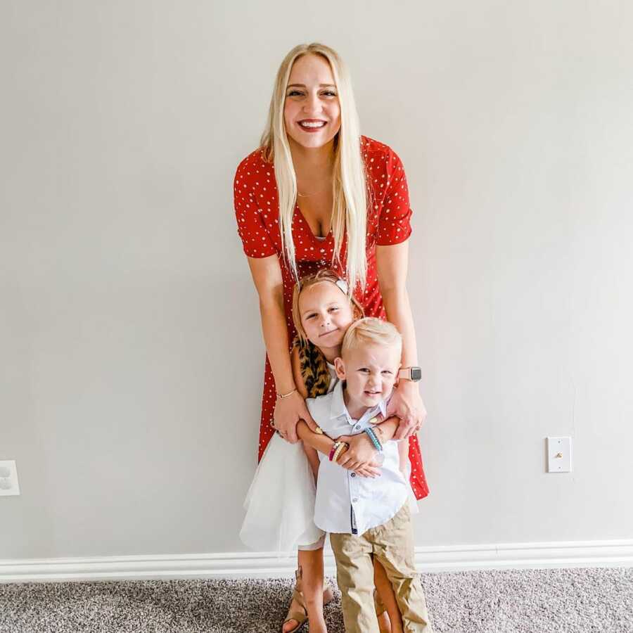 mother stands with her son and daughter in front of her while having her arms wrapped around them