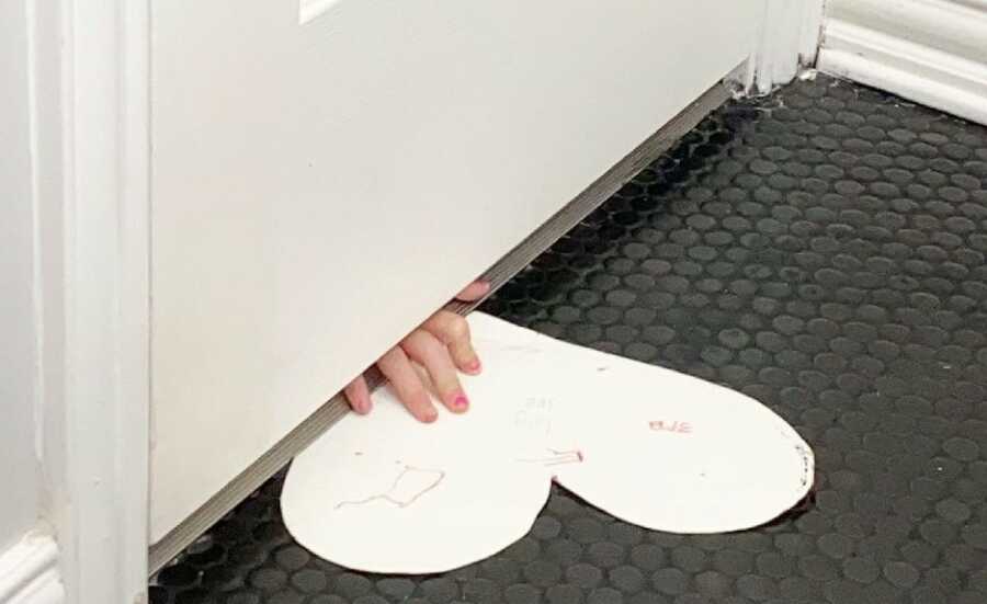 small child slides a paper heart with drawings on it under the door to show their mother