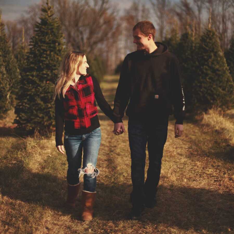 Husband and wife walking in a field of pine trees, hands interlocked as they smile lovingly at each other.