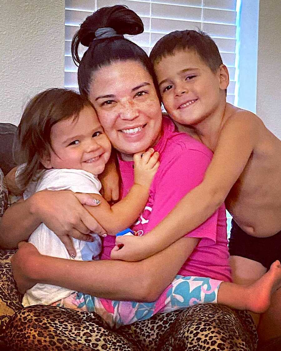 mother holds her daughter close in her lap while her son stands to the right and wraps his arm around his mother