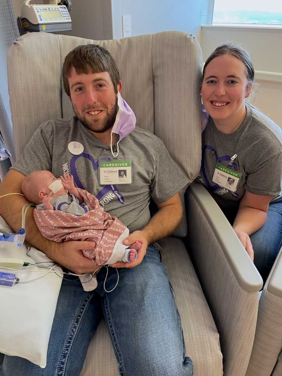 father of preemie baby girl holds her while sitting in chair while mother poses next to them