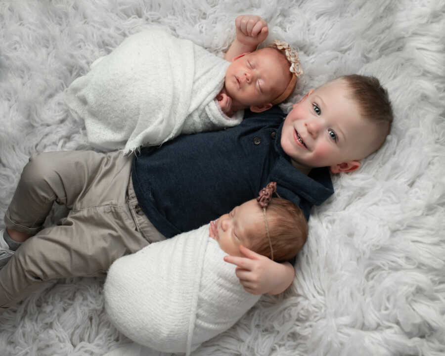Brother smiles while wrapping arms around new baby twin sisters.