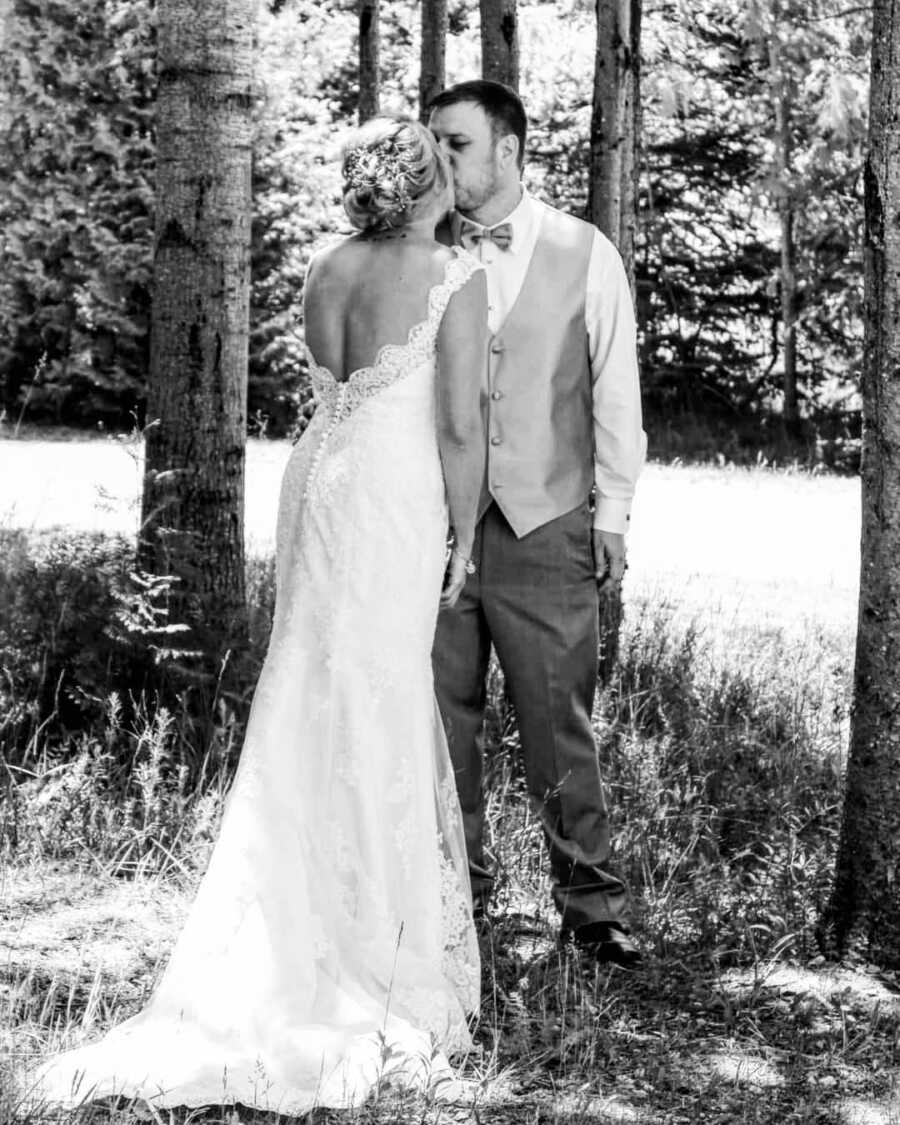 husband and wife in wedding attire posing in the forrest, wife is facing her husband and giving him a kiss. 
