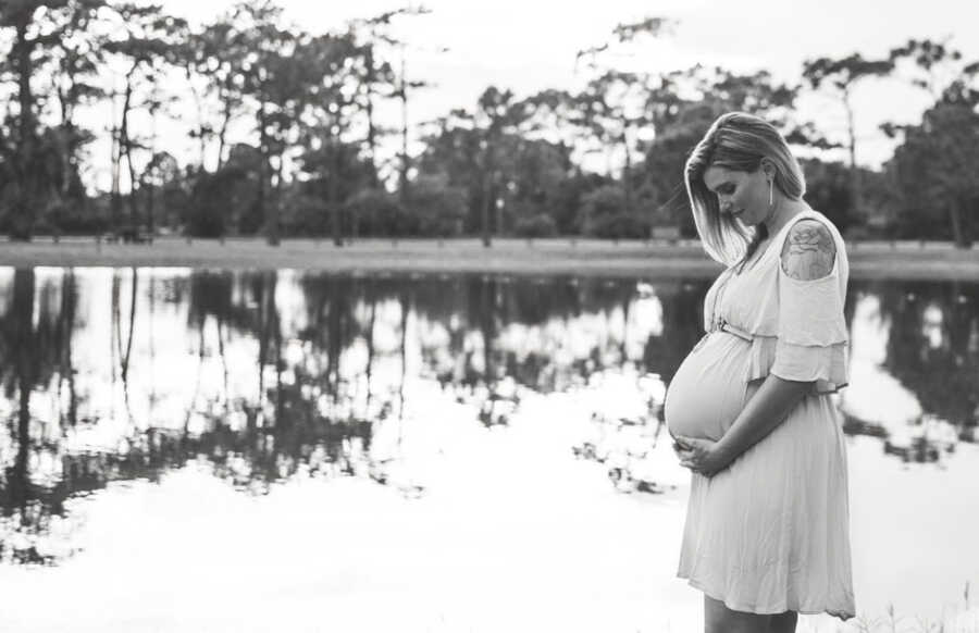 Pregnant woman holding belly with both hands wearing a dress standing in front of a lake 