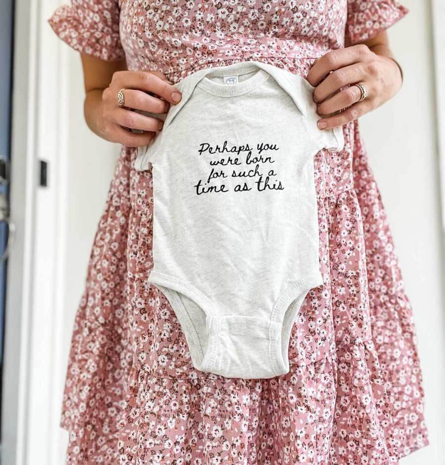 Woman pregnant with a child after three losses takes a photo of her belly bump with a onesie that reads "Perhaps you were born for a time such as this"