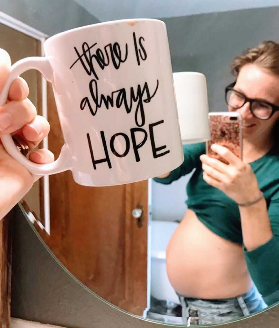 Woman pregnant after three miscarriages takes a mirror selfie of her baby bump with a mug that reads "There is always hope"
