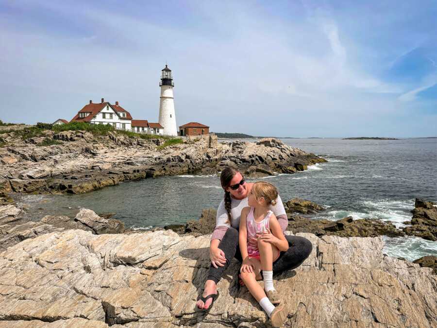 mother and daughter looking at each other while sitting on a rock at a lake in Minnesota with a light tower in the background 