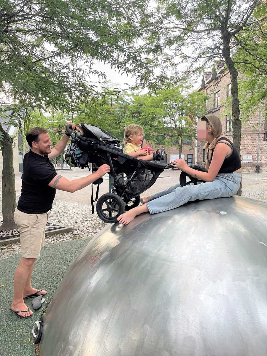 Dad holding stroller up to a giant metal ball where daughter sits playing with baby brother 