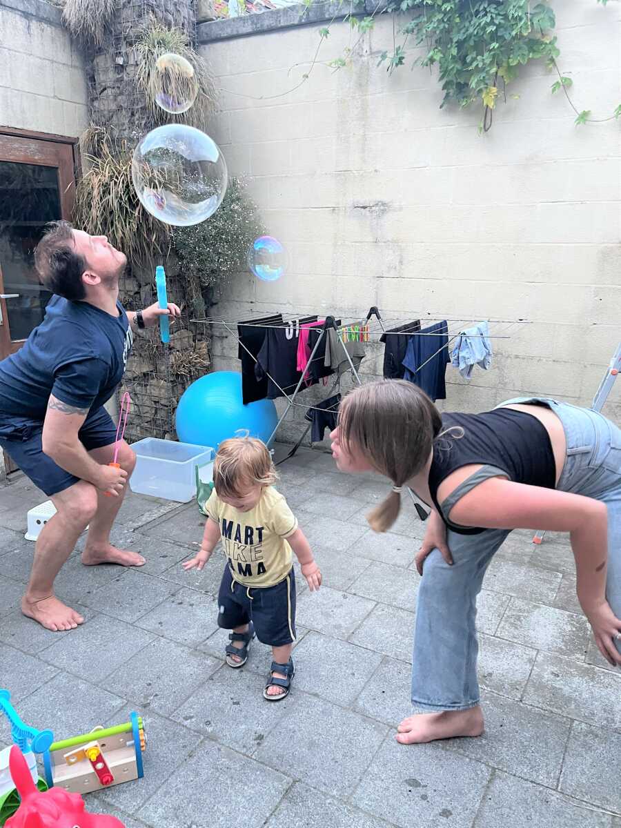 Dad blowing bubbles on the patio while teen daughter and toddle son play 