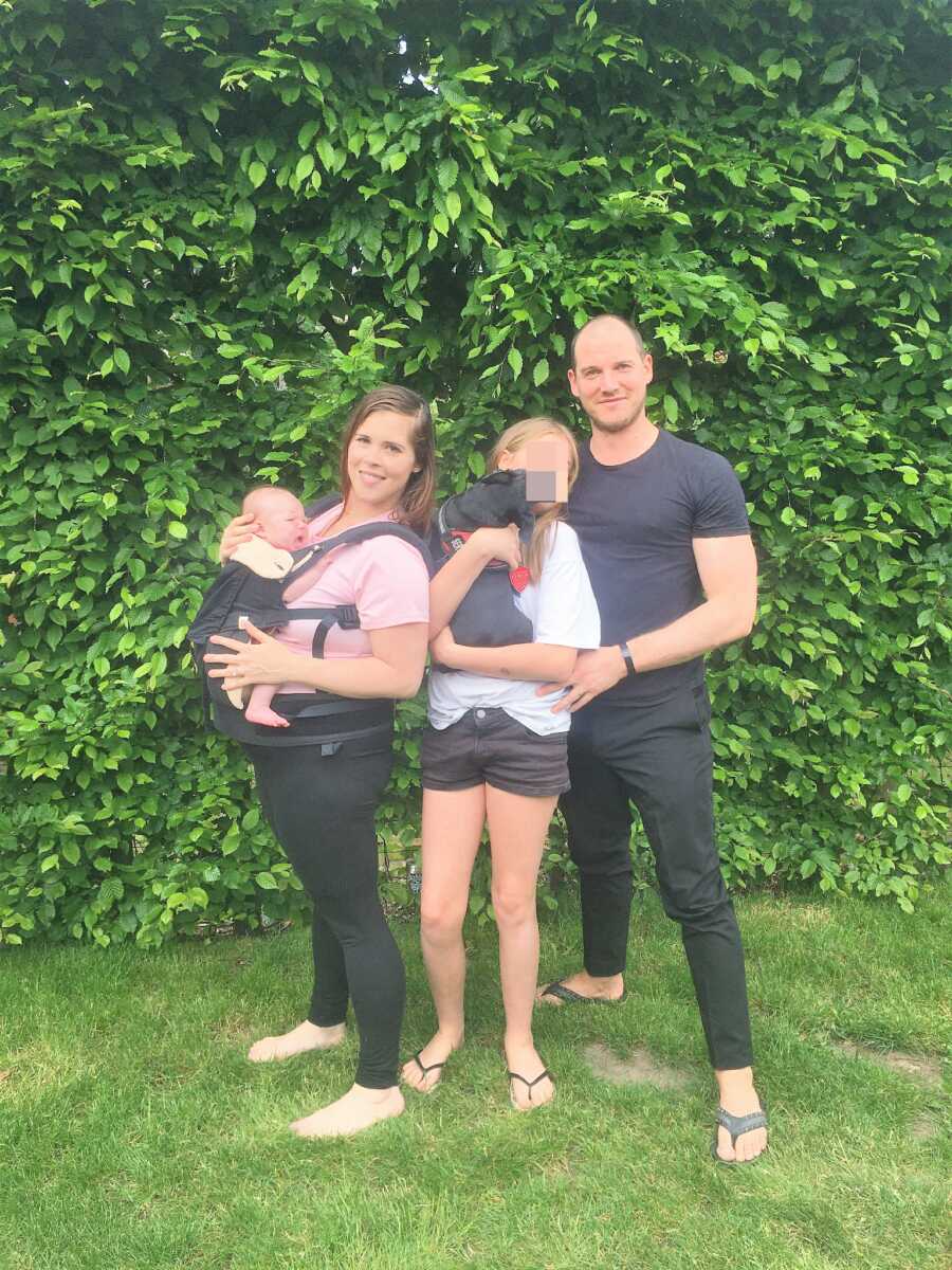 Blended family standing on grass outdoors carrying a dog and a baby 