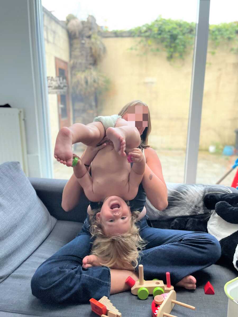preteen sister holding baby brother upside-down in the couch while they both laugh 