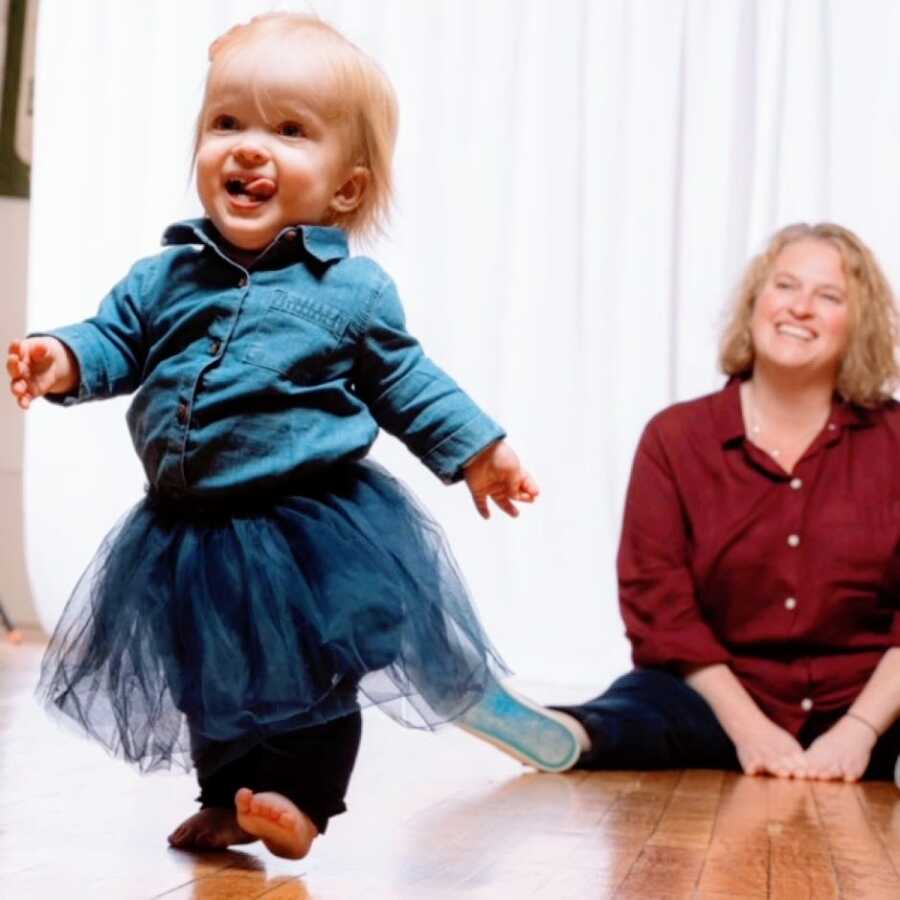 Mom in red shirt sits on the floor while her daughter runs away from her in blue shirt and tutu