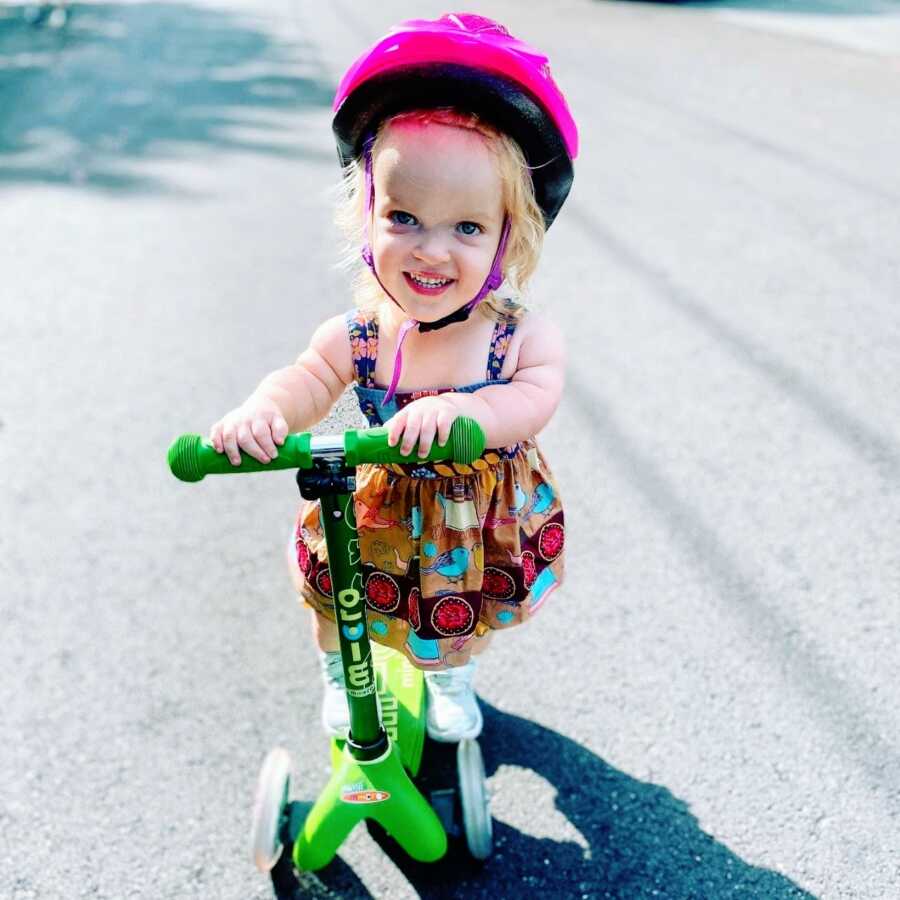 Young girl with dwarfism rides on a green scooter while wearing a pink helmet