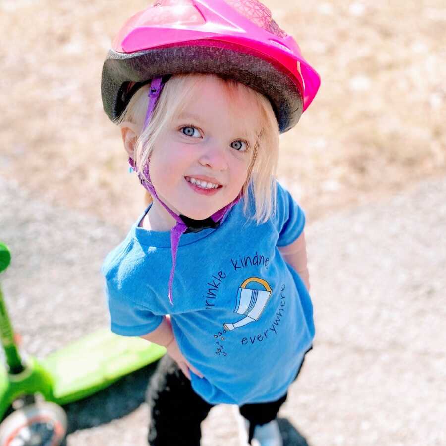 Little girl enjoying a day at the park smiles for a photo in a pink helmet while wearing a shirt about kindness