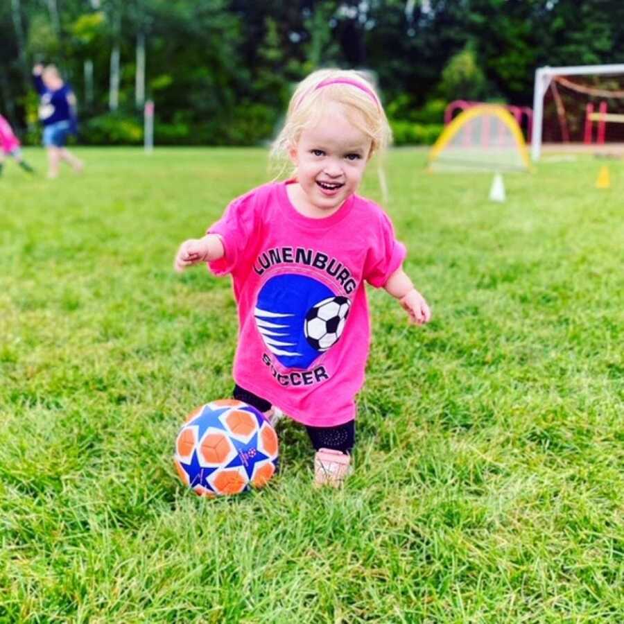 Little girl with dwarfism plays soccer on a field in a pink t-shirt